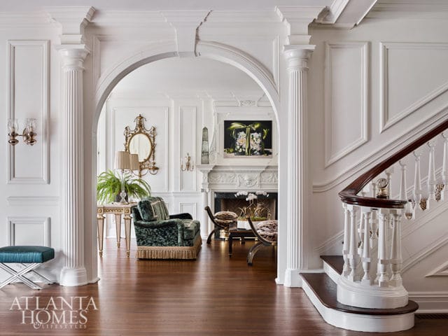 White foyer with arched doorway, fluted columns, and ornate staircase. Green velvet chair.
