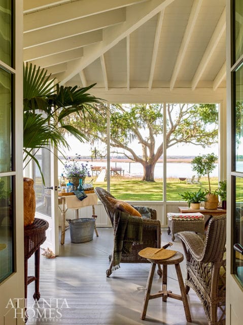 Screened porch with wicker chairs, a table with flowers, and a view of a large tree by the water