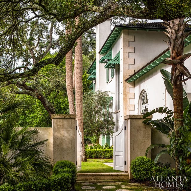 White house with green awnings and stone accents, surrounded by lush greenery and palm trees.