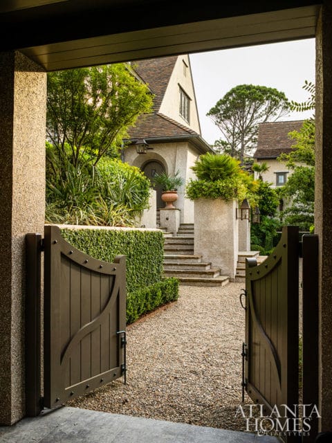 Open wooden gates leading to a stone house with lush landscaping and gravel driveway.