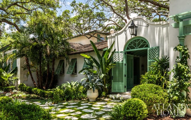White stucco house with green shutters and arched doorway, surrounded by lush landscaping.
