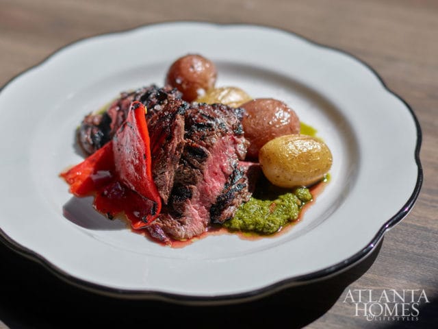 Grilled steak with roasted potatoes, red peppers, and pesto on a white scalloped plate.