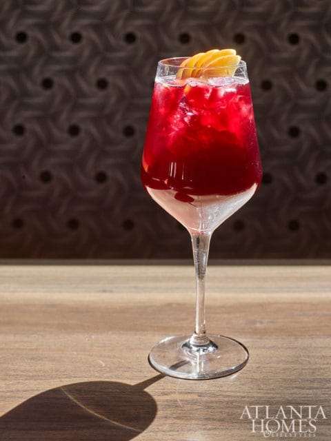 Red cocktail with ice and fruit garnish in a wine glass on a wooden table.