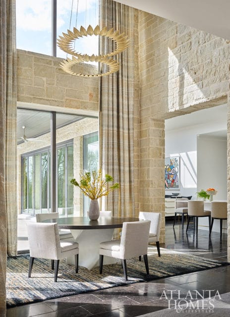 Dining room with stone walls, large windows, and a round table with upholstered chairs.