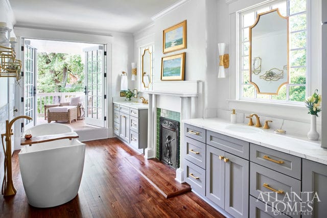 White freestanding tub with gold faucet, gray double vanity with gold hardware, and fireplace.