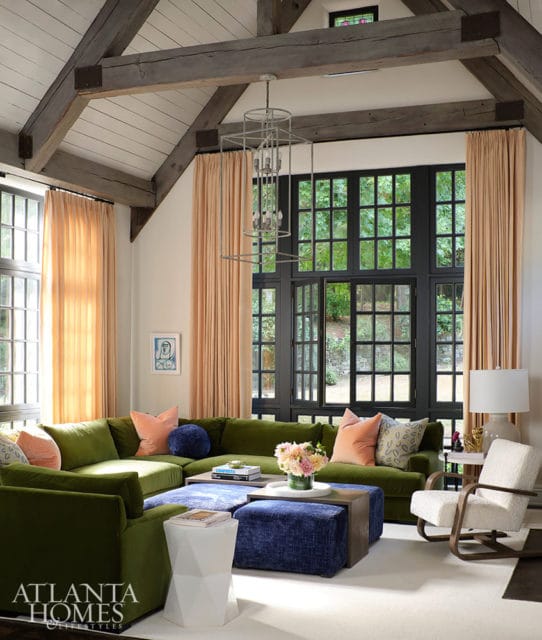 Living room with green sectional sofa, blue ottomans, and large windows overlooking trees.