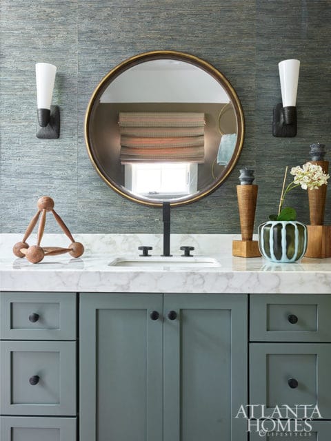 Green vanity with marble countertop, round mirror, and textured blue wallpaper.