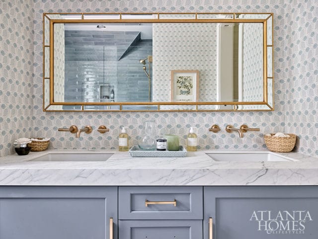 Double vanity with marble countertop, brass faucets, and blue cabinets. Patterned wallpaper and