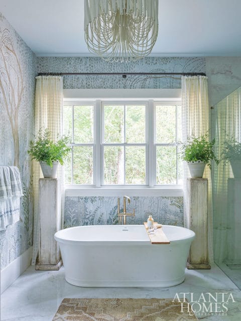 Freestanding white bathtub with brass faucet and wooden tray, under a large window.