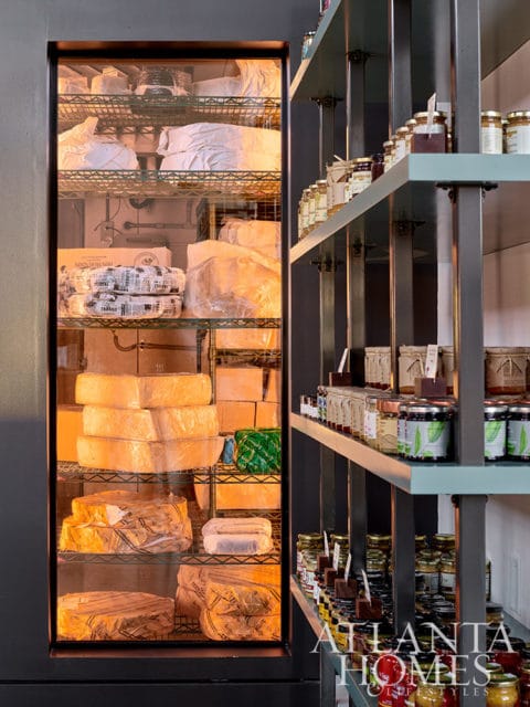 Glass-fronted refrigerator filled with cheese wheels and blocks, next to shelves of jars.