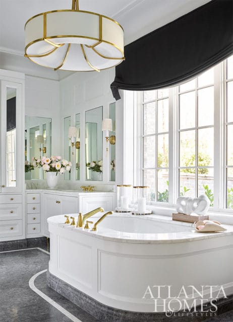 White bathroom with freestanding tub, marble countertops, brass fixtures, and large windows.