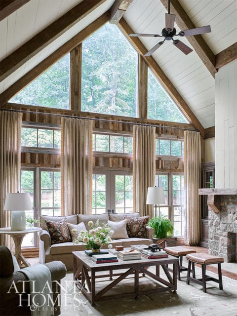Living room with vaulted wood-beamed ceiling, large windows, stone fireplace, and neutral furniture.