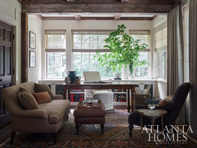 Study with taupe sofa, leather ottoman, dark armchair, and wooden desk by bay windows.