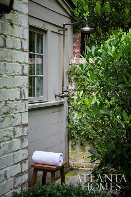 Outdoor shower with brass fixtures next to a gray-shingled building and lush greenery.