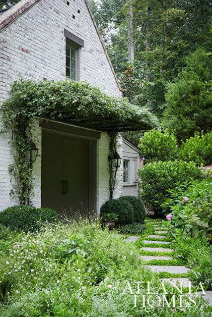 White brick building with large double doors, covered by a lush green vine canopy. Stone path leads