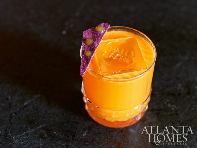 Orange cocktail with ice cube and purple garnish, Atlanta Homes & Lifestyles logo