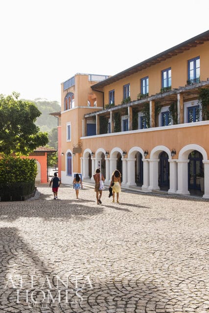 Orange building with arched colonnade and blue windows, people walking on cobblestone path.