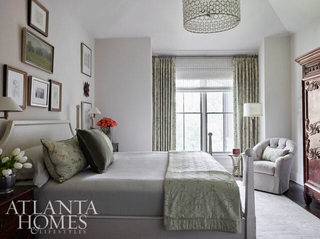 Bedroom with a white upholstered headboard, grey bedding, and a green embroidered throw. Floral curtains frame a large window, and a tufted armchair sits beside an ornate armoire.