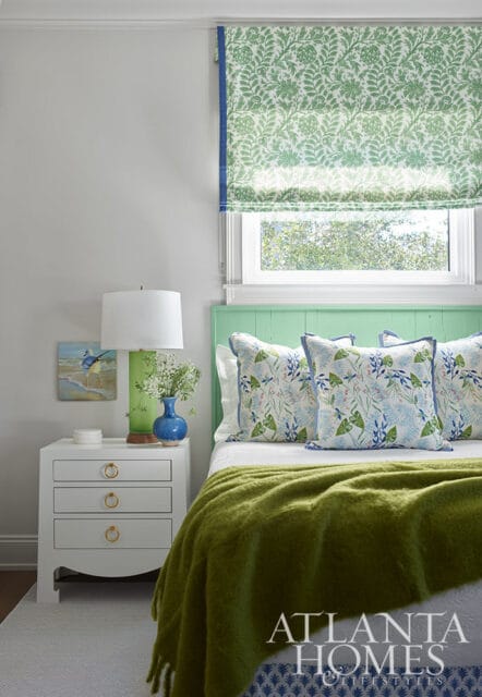 Bedroom with green headboard, floral pillows, green throw, and patterned roman shade.