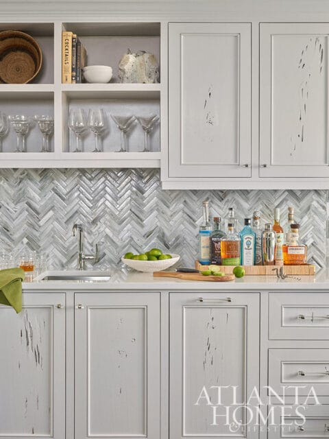 Gray bar cabinet with herringbone tile backsplash, sink, and bottles of liquor.