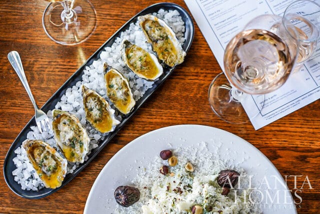 Six baked oysters on a black platter filled with crushed ice.