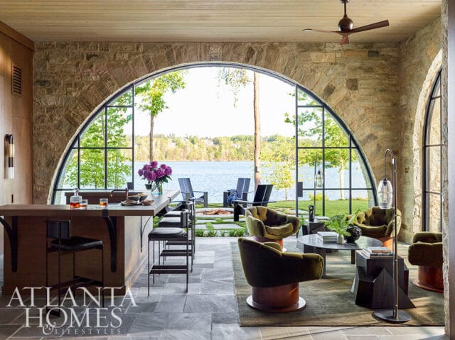 Living room with stone arch window overlooking lake, wood bar, and green velvet chairs.