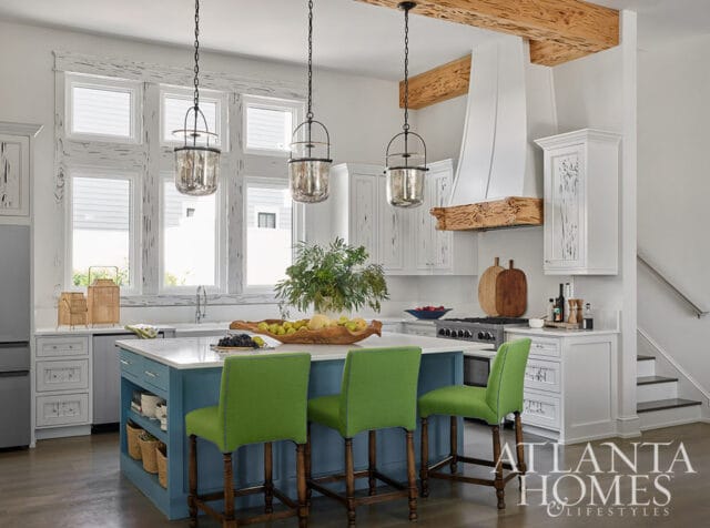 Blue kitchen island with green bar stools, white cabinets, and large windows.