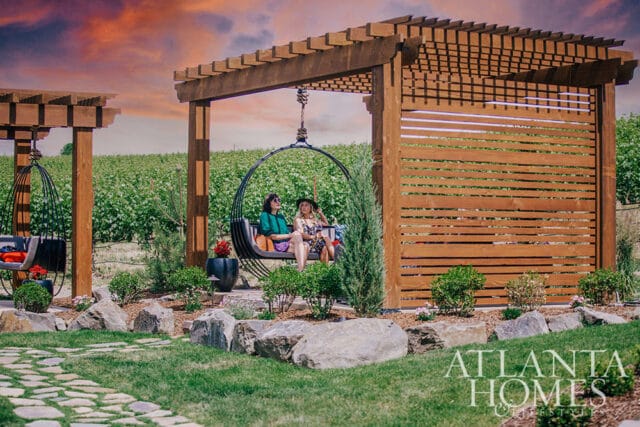 Two women sit in hanging chairs under a wooden pergola overlooking a vineyard at sunset.