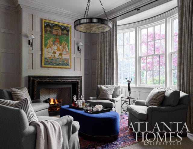 Living room with gray armchairs, blue ottoman, fireplace, and artwork above mantel.