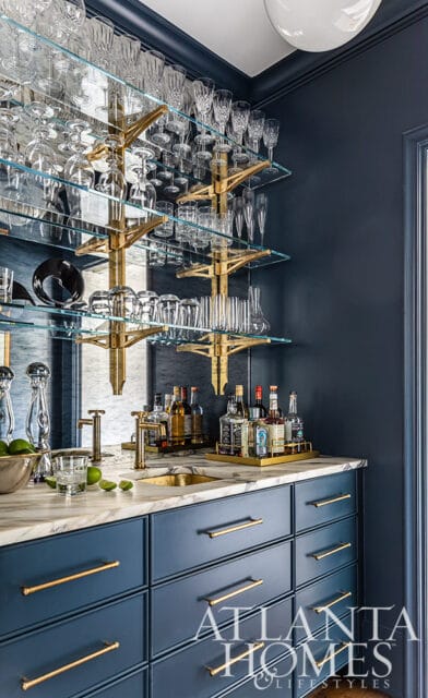 Bar with navy cabinets, marble countertop, brass fixtures, and glass shelves of glassware.