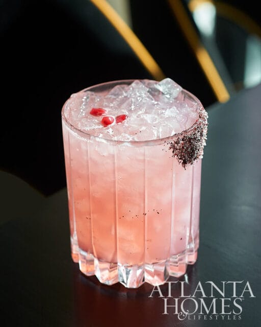 Pink cocktail with ice, pomegranate seeds, and a dark rim garnish in a fluted glass.