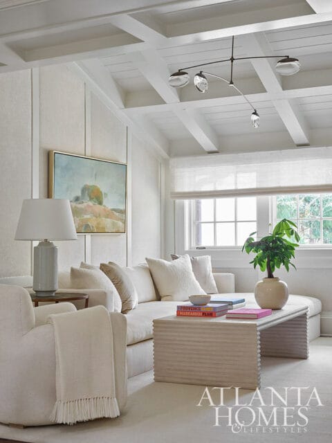 Living room with white sectional sofa, textured armchair, coffered ceiling, and abstract art.