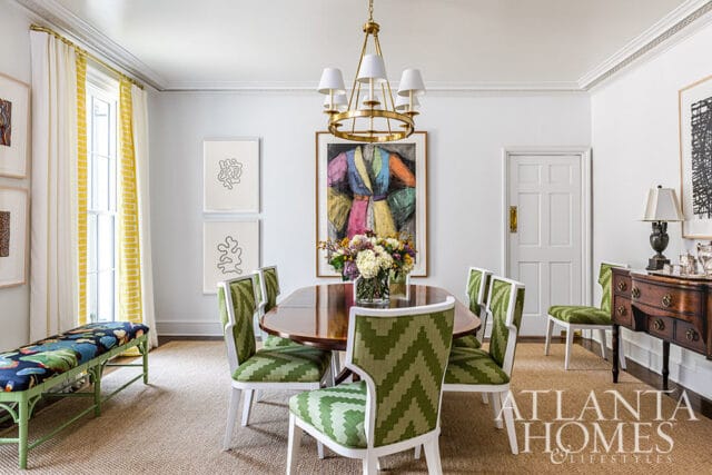 Dining room with oval wood table, green chevron chairs, colorful art, and floral centerpiece.