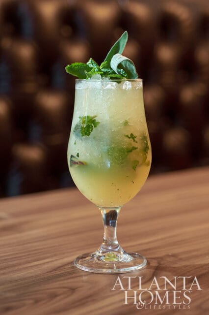 Yellow cocktail with mint and sage garnish in a stemmed glass on a wooden table.