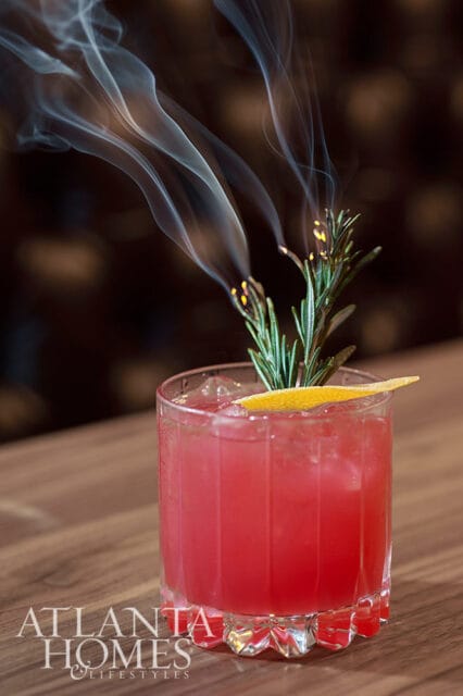 Pink cocktail with ice, orange peel, and smoking rosemary sprig