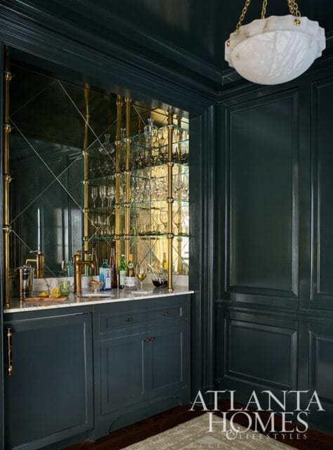 Dark green bar with marble countertop, mirrored backsplash, and brass shelving stocked with