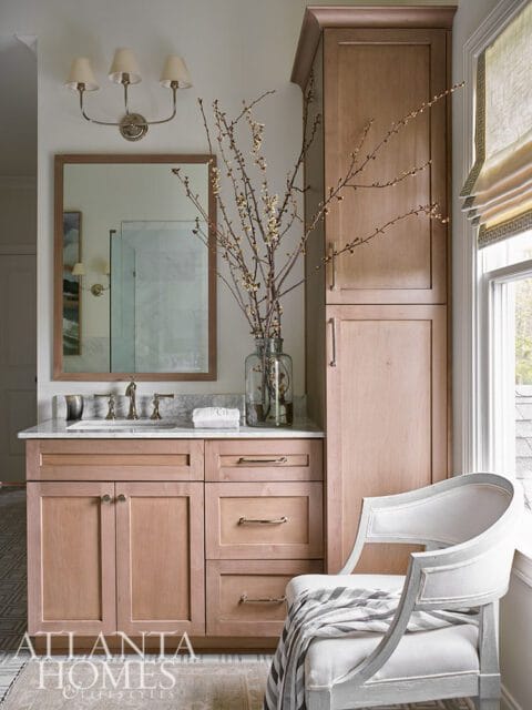 Wood vanity with marble countertop, mirror, and tall cabinet. White chair with striped blanket.