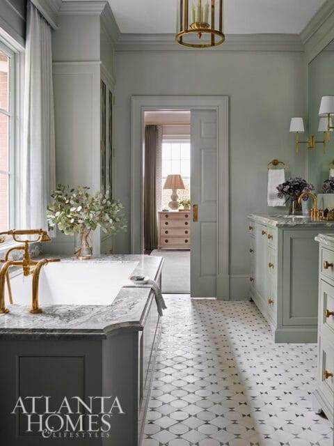 Sage green bathroom with marble tub surround, brass fixtures, and patterned tile floor.