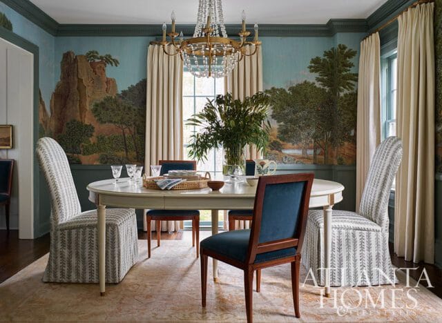 Dining room with a large oval table, upholstered chairs in patterned fabric and blue velvet, and a scenic mural wallpaper depicting mountains and trees.