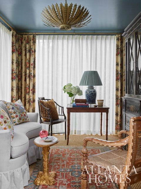 Blue ceiling, floral curtains, and a gold fan chandelier over a table with a blue and white lamp.