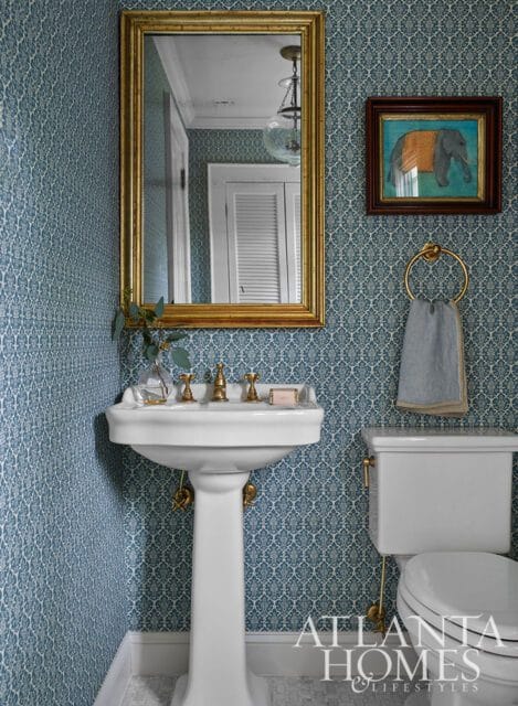 Powder room with blue patterned wallpaper, white pedestal sink, brass fixtures, and gold framed