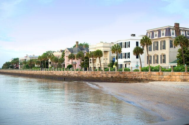 Row of historic homes with palm trees along a waterfront promenade and sandy beach.