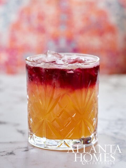 Two-toned cocktail with ice in a faceted glass, layered with orange and red liquids, on a marble surface.
