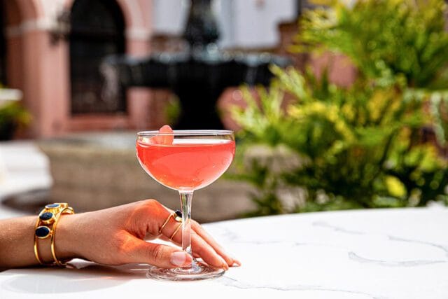 Pink cocktail in coupe glass with watermelon garnish, hand with jewelry resting on table