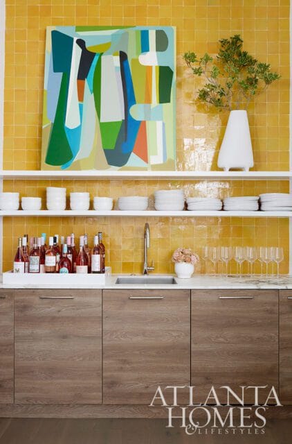 Yellow tiled backsplash with abstract art above a shelf of white dishes and bottles of wine.