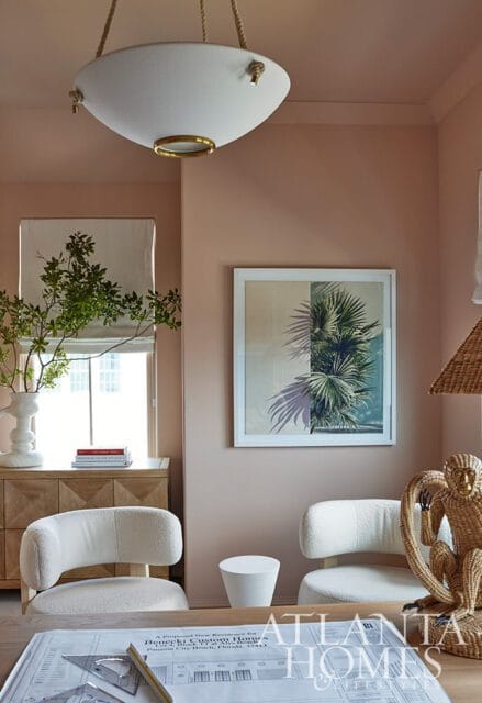 Pink walls, white bouclé chairs, framed palm leaf art, and a woven monkey statue.