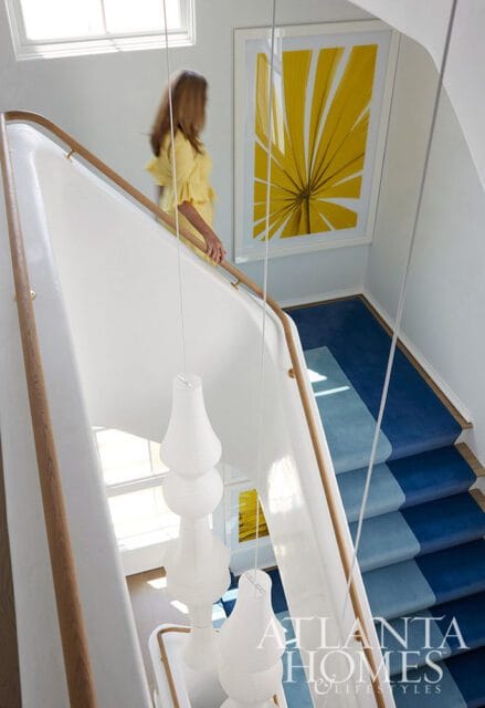 White staircase with wooden handrails, blue ombre carpet runner, and yellow palm leaf art.
