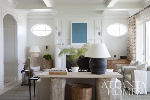 Living room with white paneled walls, oval windows, fireplace, and light neutral furniture.