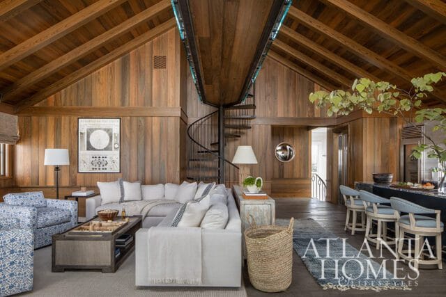 Living room with vaulted wood-paneled ceiling, spiral staircase, sectional sofa, patterned armchairs, and a kitchen island with bar stools.