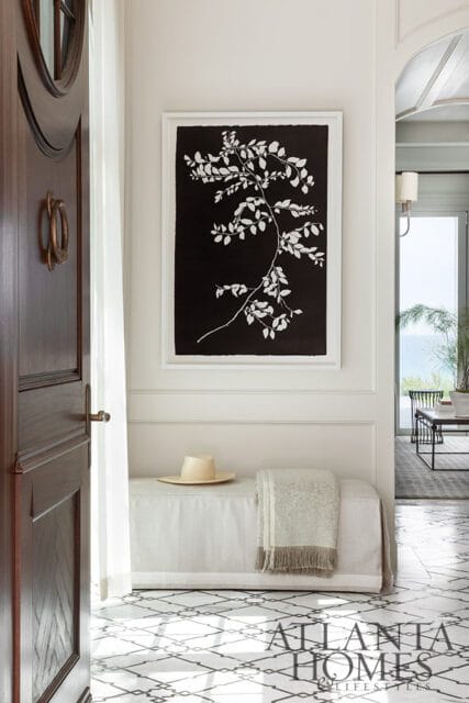 Foyer with dark wood door, white bench with hat and blanket, and framed botanical art.